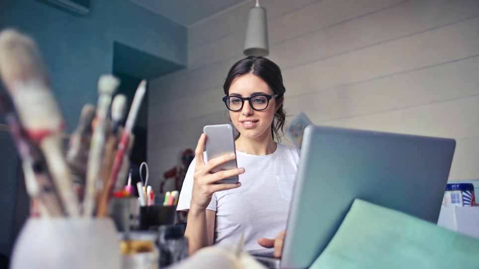 Woman in studio with paint brushes and laptop in the foreground looking at her smart phone with a smile